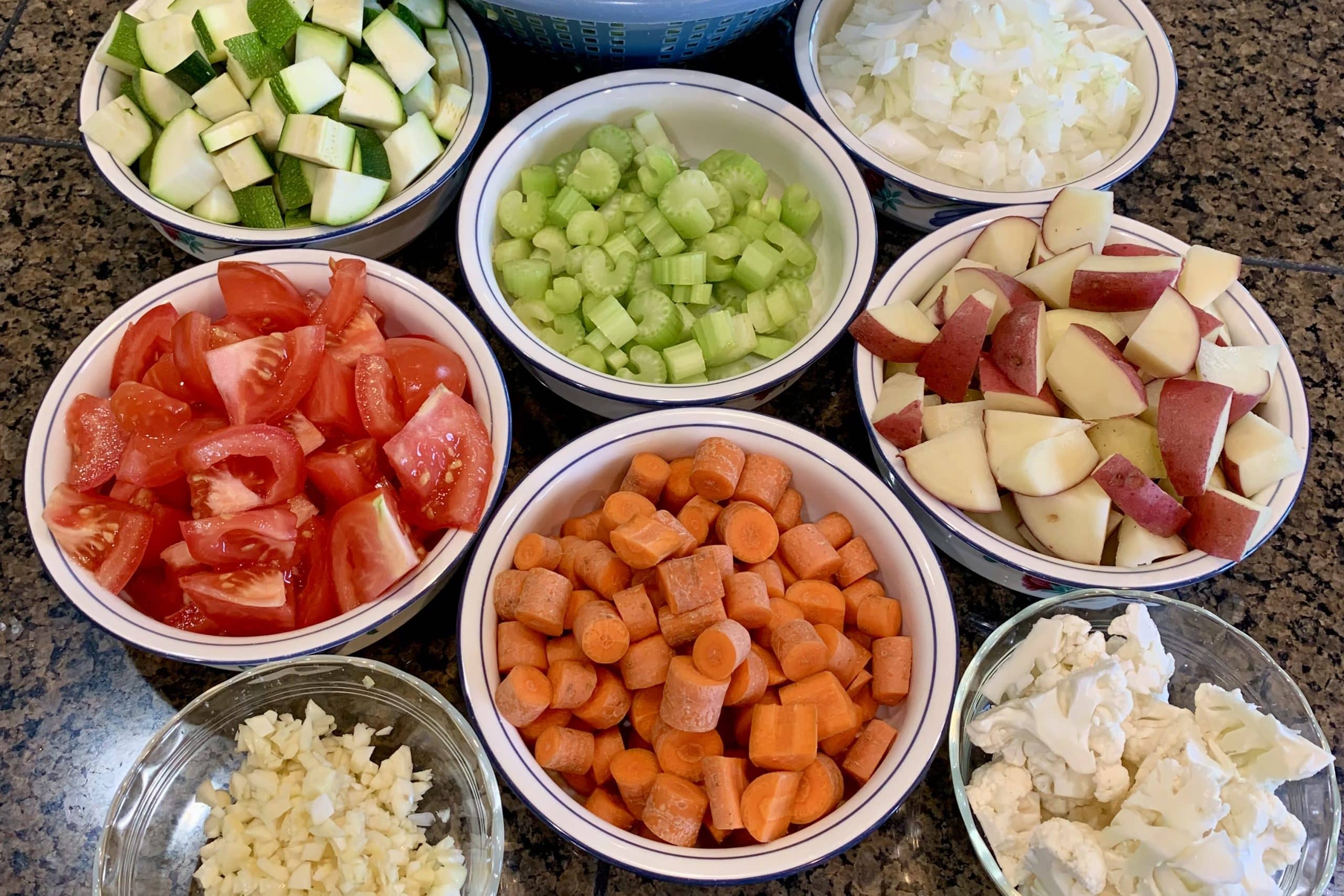 Fresh chopped vegetables for healthy cooking: diced zucchini, chopped onions, celery chunks, quartered tomatoes, sliced carrots, cubed red potatoes, minced garlic, and cauliflower florets arranged in separate white bowls on a granite countertop. Plant-based meal prep ingredients for longevity and wellness recipes.