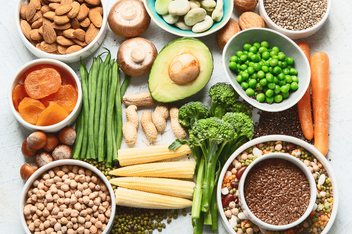 Flat lay of assorted plant-based protein sources including chickpeas, lentils, green peas, avocado, almonds, hazelnuts, carrots, mushrooms, broccoli, flax seeds, and whole grains on a light background.