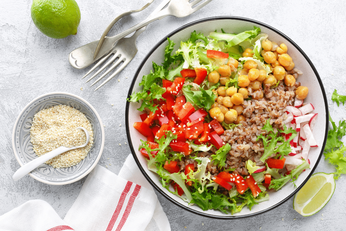 Vibrant plant-based protein bowl with chickpeas, brown rice, leafy greens, radishes, and red bell peppers topped with sesame seeds, served with a side of sesame and lime.