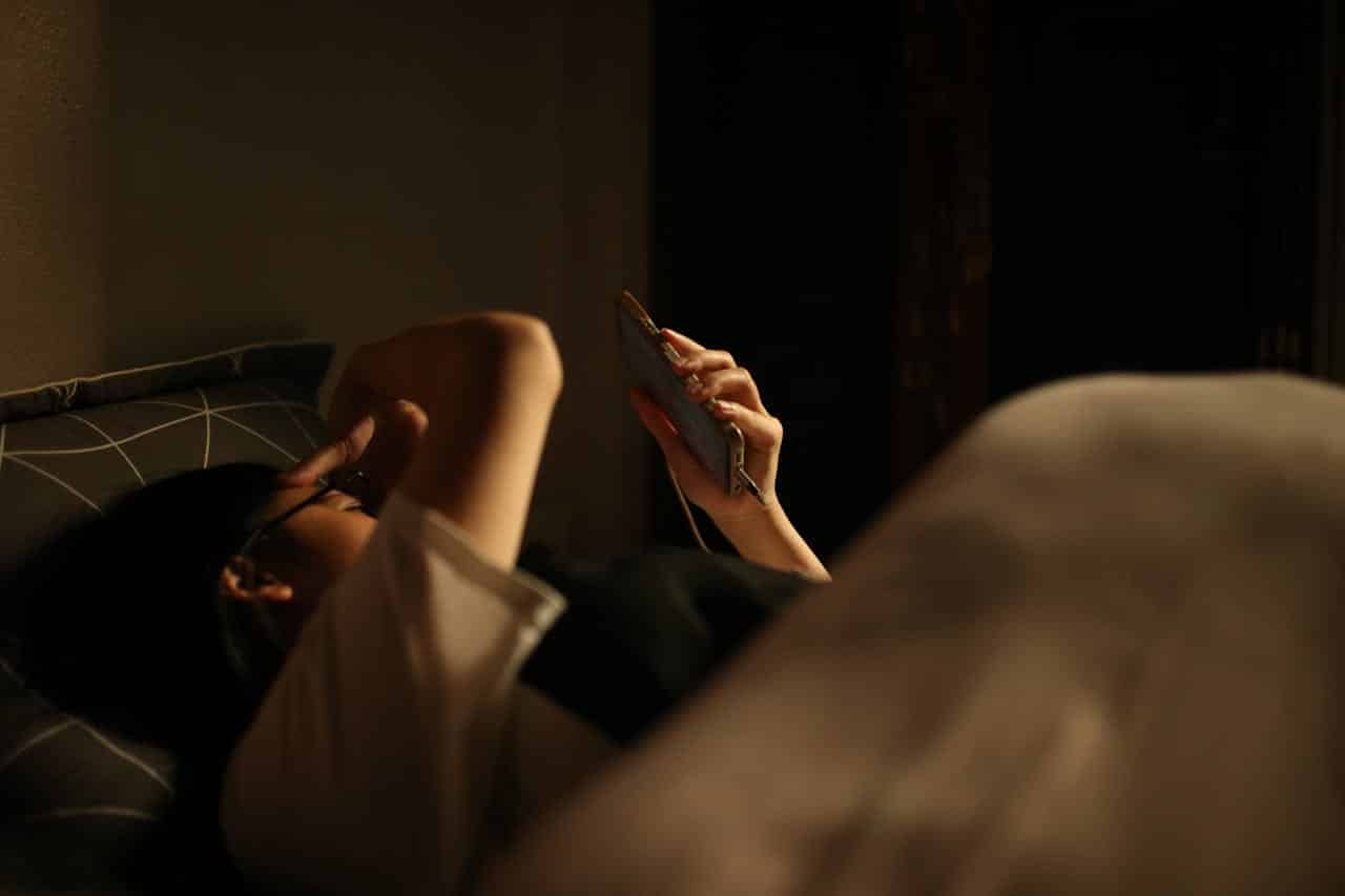 Person using phone in dark bedroom, demonstrating harmful blue light exposure that disrupts natural sleep cycles and longevity. The nighttime screen use illustrates how modern technology can impact sleep quality and overall lifespan