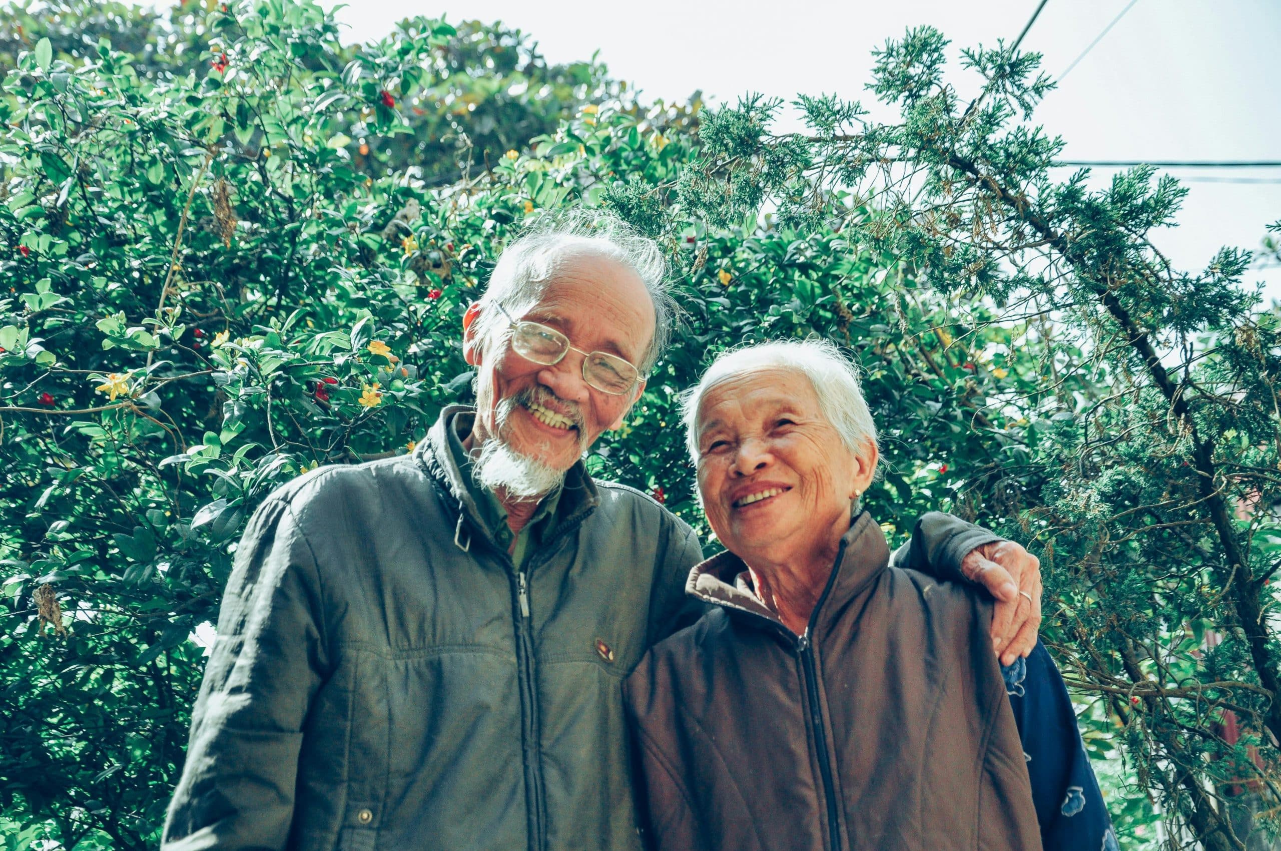A joyful elderly Asian couple smiling at the camera while standing in a lush garden. They're both wearing casual outdoor jackets, and the man has glasses and a white beard. The natural green foliage background with small red flowers suggests a healthy, active lifestyle. The image conveys vitality and wellness in aging, perfect for illustrating content about longevity and healthy dietary choices