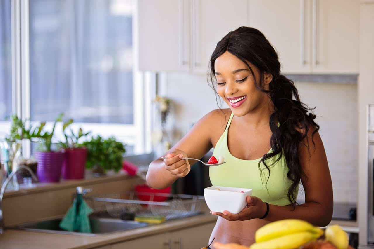 Young woman smiling while enjoying healthy breakfast in bright kitchen, wearing activewear and eating fresh fruit from white bowl. Windowsill herbs and bananas visible on counter, suggesting healthy plant-based lifestyle and wellness focus