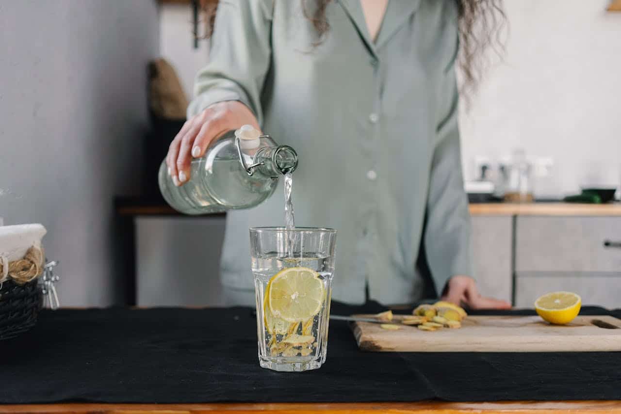 Person pouring alkaline water into a glass with fresh lemon slices, demonstrating an anti-inflammatory morning ritual for longevity. Daily wellness practice shown in modern kitchen, featuring citrus-infused water for reduced inflammation, improved digestion, and cellular health. Part of an anti-aging hydration routine with antioxidant-rich lemon water for optimal wellness and immune support