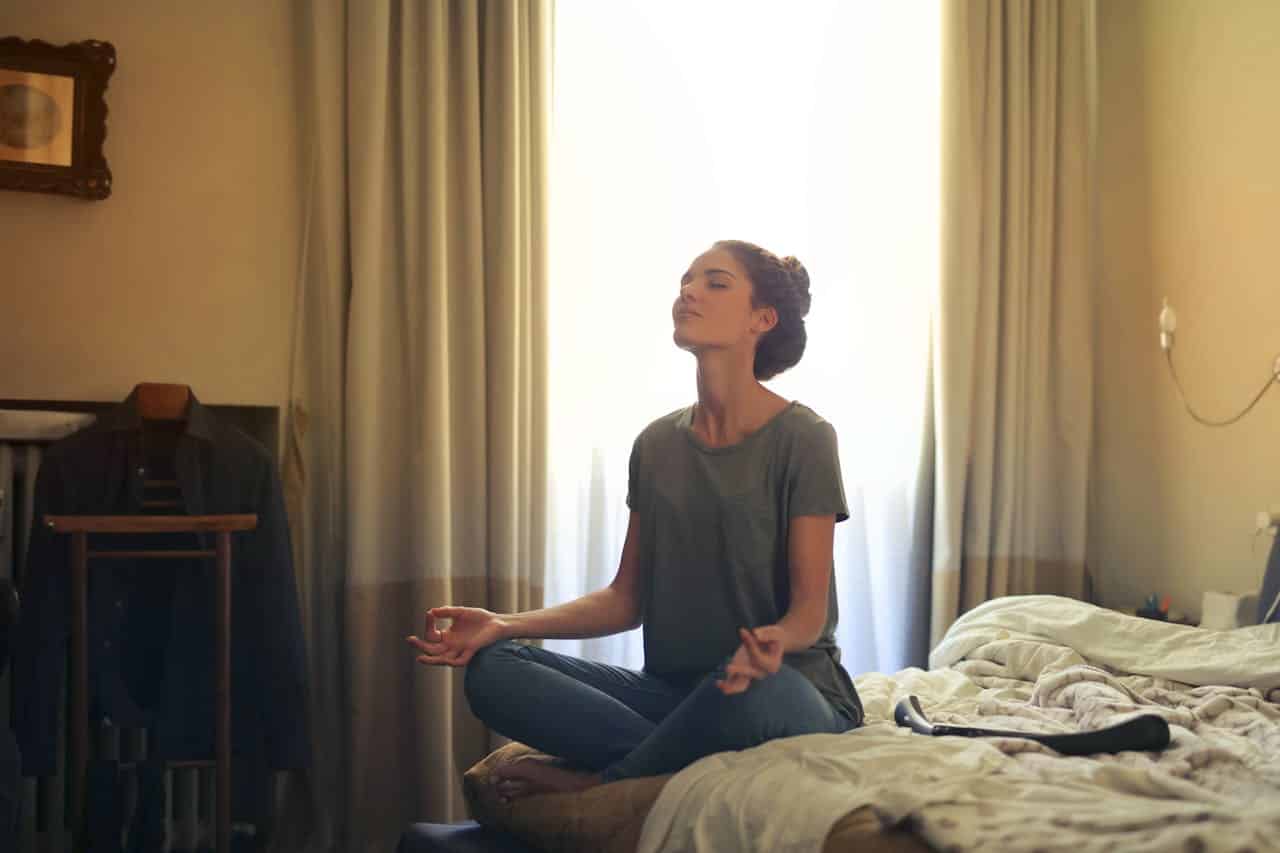 Person meditating on bed in morning light, demonstrating mindful relaxation techniques for better sleep and longevity. The peaceful bedroom setting highlights the connection between evening meditation, quality rest, and a longer, healthier life