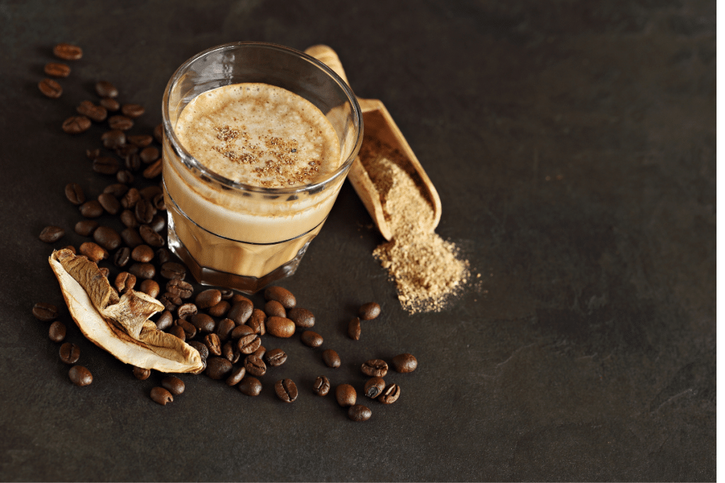A creamy glass of mushroom coffee on a dark stone surface, surrounded by coffee beans, dried mushrooms, and a wooden scoop of mushroom powder.