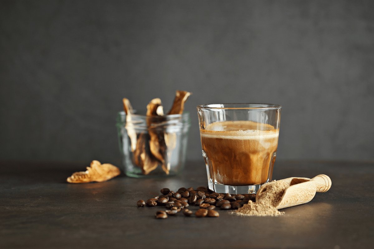 Glass of mushroom coffee with visible crema, surrounded by coffee beans, dried mushrooms in a glass jar, and a wooden scoop of mushroom powder on a dark rustic background.