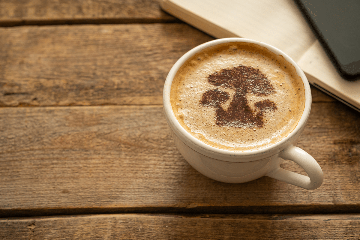 A cup of mushroom coffee on a rustic wooden table with a mushroom-shaped design in the foam, next to an open notebook and smartphone.