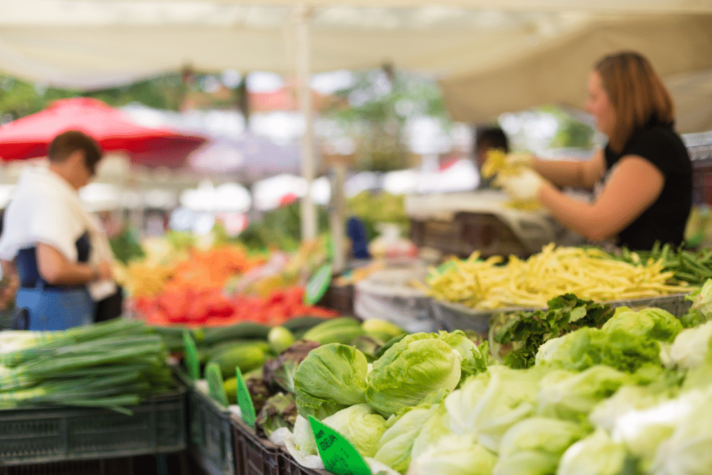 A vibrant Mediterranean market with fresh green lettuce, colorful vegetables, and people shopping under shaded stalls.