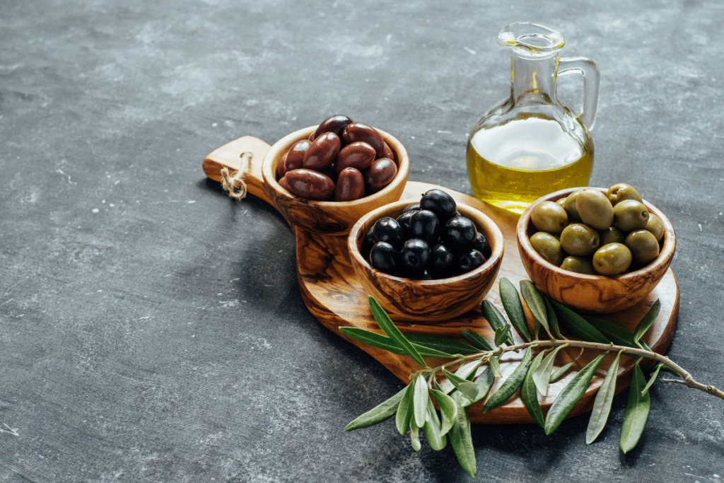 A wooden board with bowls of black, green, and Kalamata olives alongside a glass bottle of extra virgin olive oil and fresh olive branches.