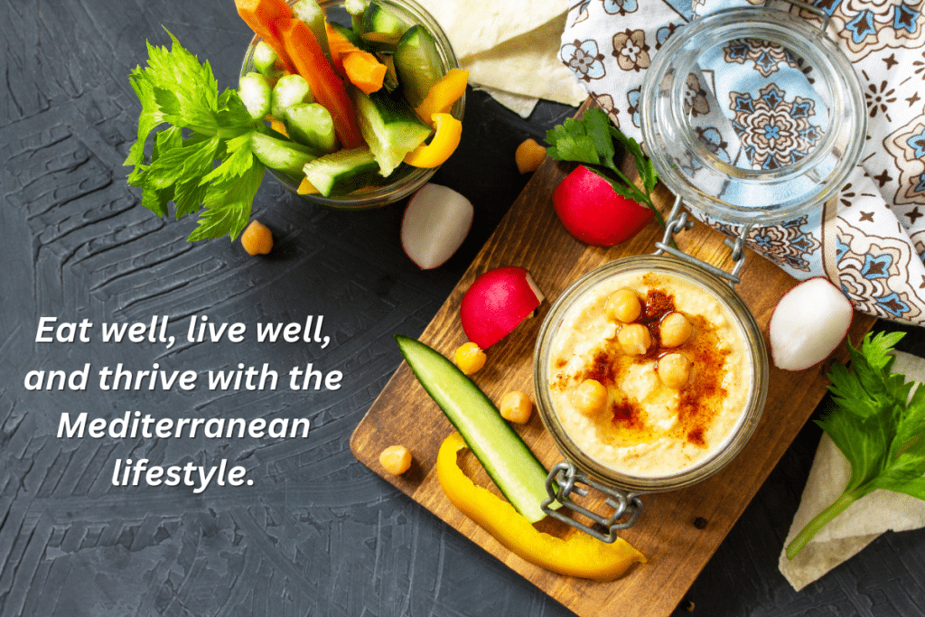 Overhead view of Mediterranean-style snacks featuring homemade hummus topped with chickpeas and paprika in a glass jar, alongside fresh cut vegetables including cucumber, yellow bell pepper, celery, and radishes on a wooden serving board. A patterned cloth napkin and empty glass jar complete the rustic presentation on a dark slate surface.