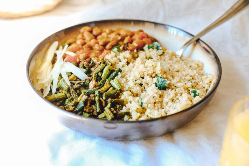 Healthy grain bowl featuring quinoa, sautéed green vegetables, beans, and fresh sprouts arranged in sections in a metallic bowl