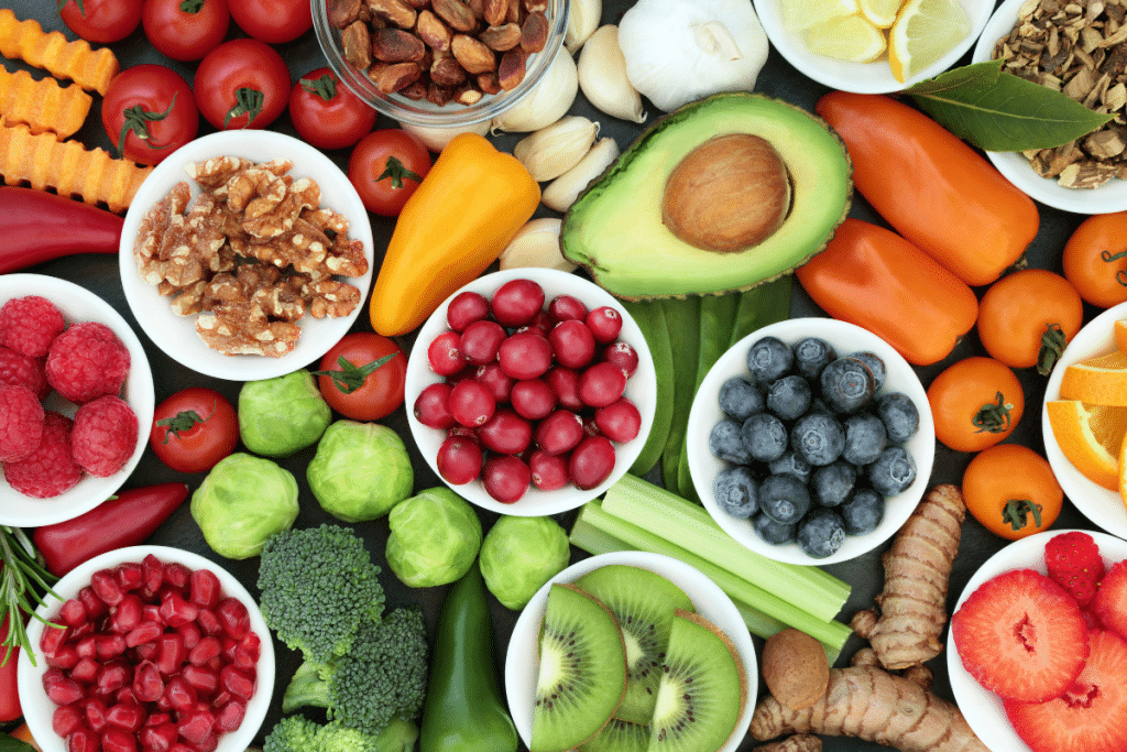 A colorful array of fresh, nutrient-rich foods arranged on a dark background. The spread includes raspberries, blueberries, tomatoes, walnuts, garlic, avocado, ginger, citrus fruits, broccoli, bell peppers, cranberries, kiwi, celery, and various nuts. Foods are presented in white bowls or naturally displayed, creating a vibrant mosaic of healthy ingredients.