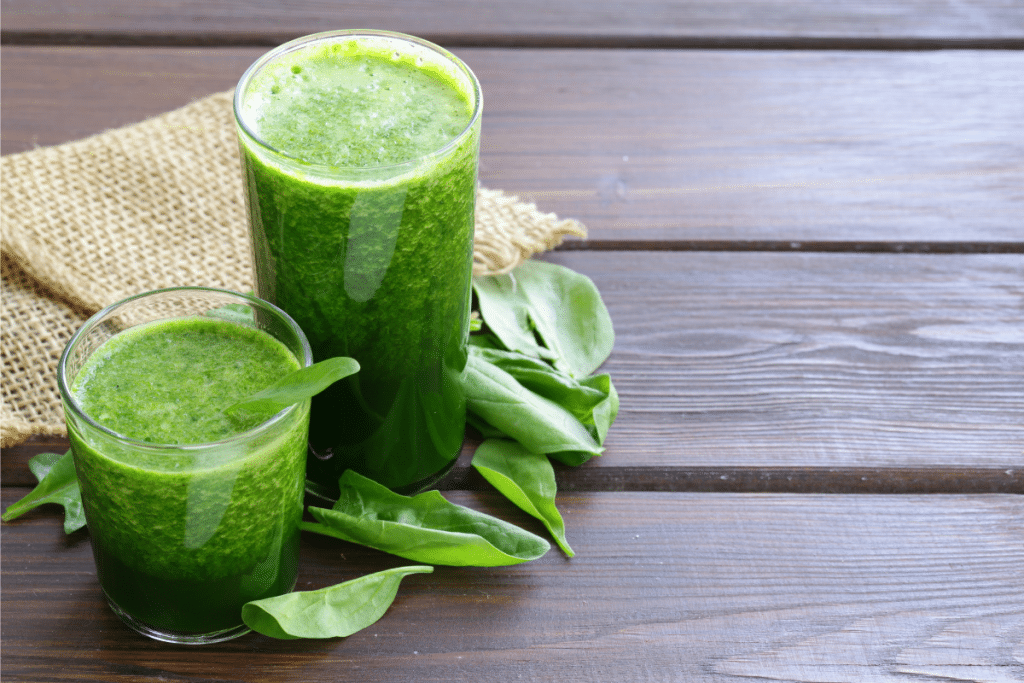 Fresh green smoothie made with leafy greens and nutrient-rich ingredients in two glass cups on wooden surface with raw spinach leaves