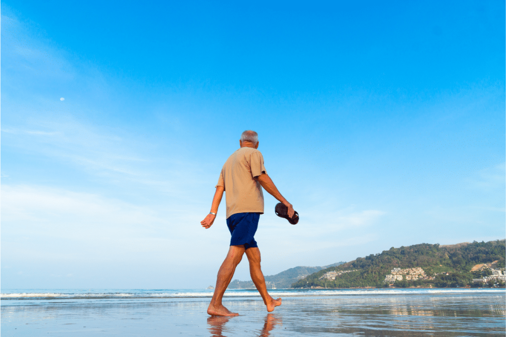 Older adult walking barefoot on beach during morning exercise, demonstrating active aging and natural movement for longevity