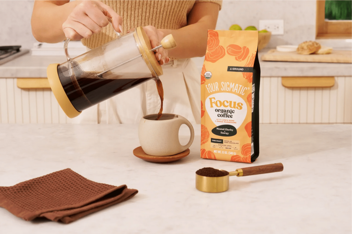 Pouring Four Sigmatic Focus coffee from French press into ceramic cup, with organic coffee bag and measuring scoop on marble countertop, demonstrating mindful morning ritual