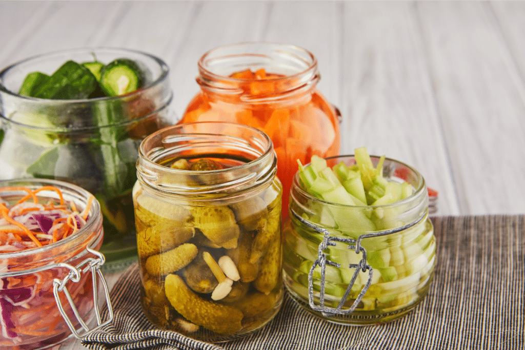 Variety of homemade fermented vegetables in glass jars including pickles, carrots, cucumber slices, and colorful cabbage slaw for gut health