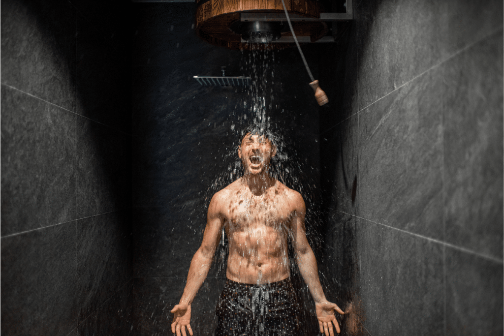 A shirtless person stands under a rainfall showerhead taking a cold shower, with an intense facial expression showing reaction to the cold water. Dark slate walls frame the dramatic scene as water droplets spray and cascade around them, demonstrating cold exposure therapy.