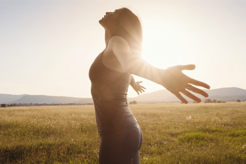 A woman standing in an open field with her arms outstretched, embracing the sunlight. The golden rays shine through her silhouette, symbolizing the health benefits of natural sunlight exposure.