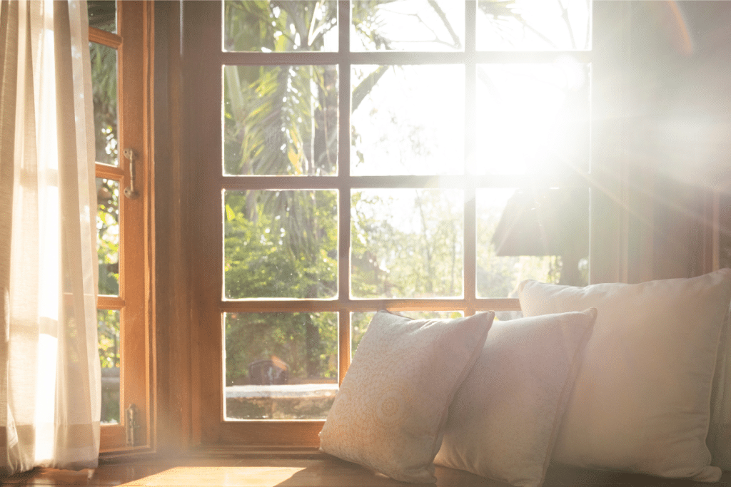 A cozy indoor space with sunlight streaming through a wooden-framed window, illuminating soft pillows on a windowsill. The warm glow highlights the connection between natural light and sleep quality.