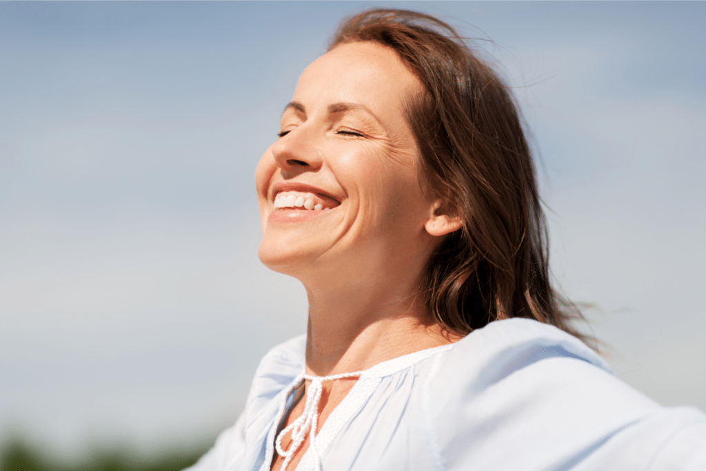 A smiling woman with her face tilted toward the sun, eyes closed, enjoying the warmth and natural benefits of sunlight. The bright sky in the background symbolizes the connection between sunlight and heart health.