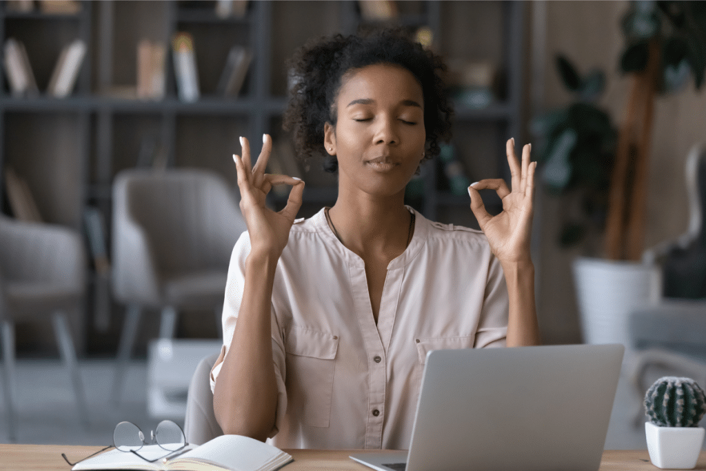 A calm woman sitting at her desk with eyes closed, meditating and practicing mindfulness to reduce stress.