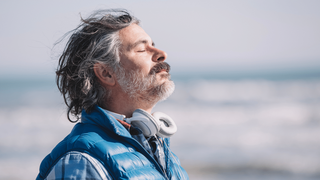 Middle-aged man with eyes closed, breathing deeply by the ocean, practicing breath work for stress relief and longevity.