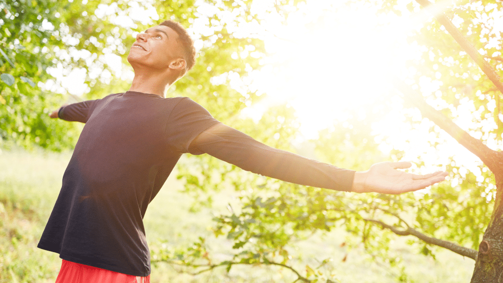 Man standing in nature with arms outstretched, smiling into the sunlight, feeling energized, refreshed, and alive.